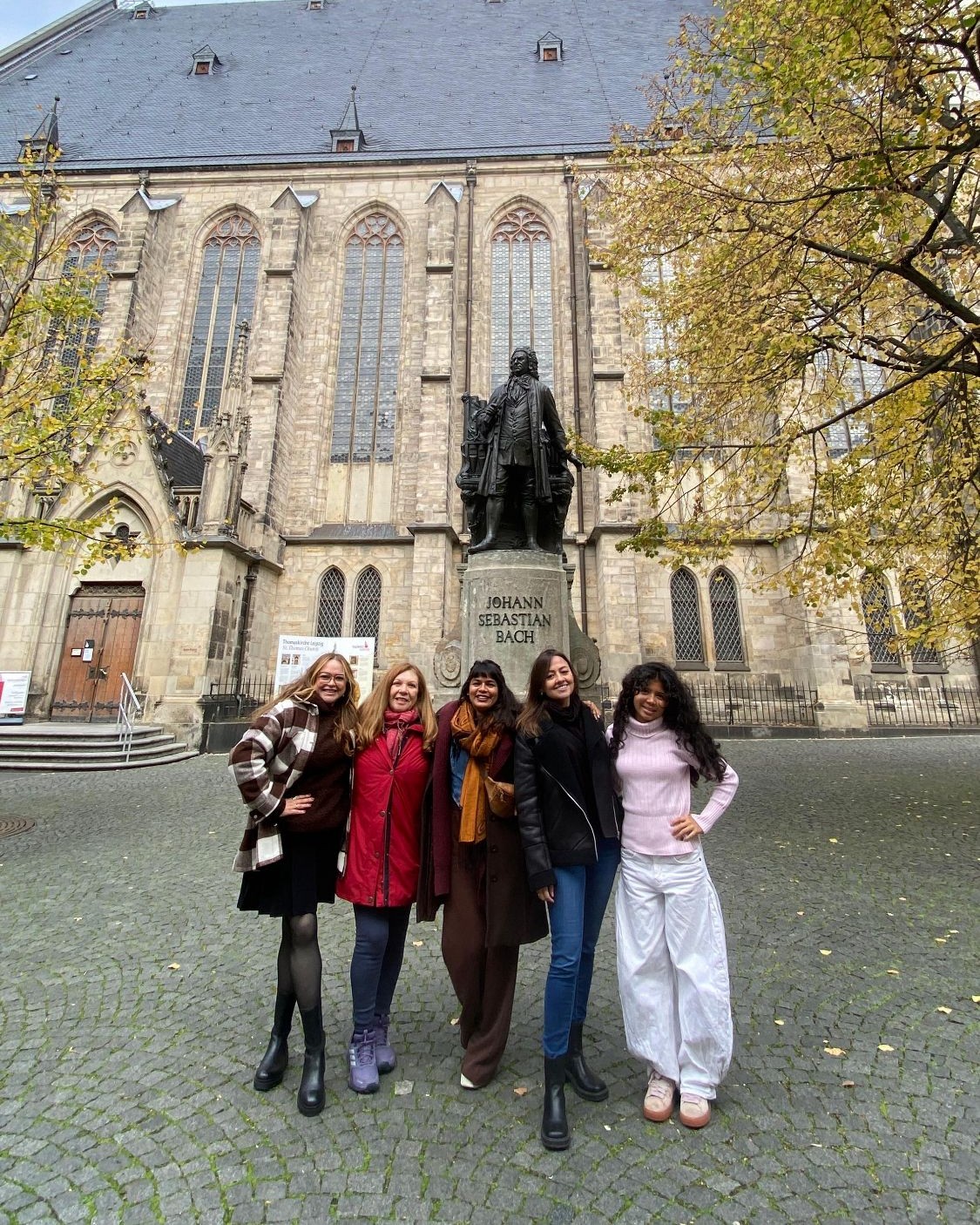 Cinco mulheres sorridentes posando em uma praça de paralelepípedos em frente a uma estátua escura de Johann Sebastian Bach, com uma antiga igreja de pedra ao fundo.