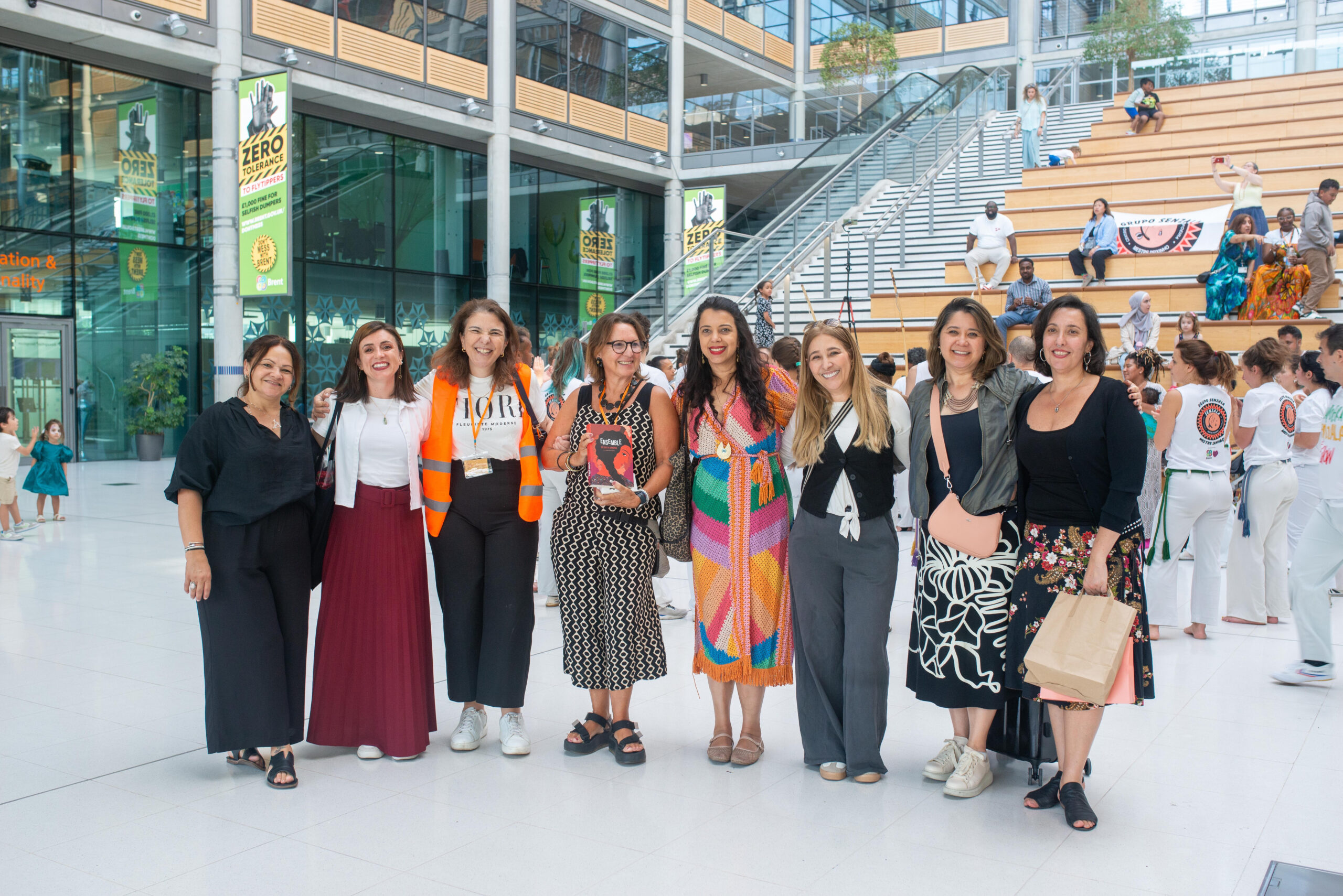 Oito mulheres sorrindo, posando em linha em um grande átrio moderno e envidraçado. O fundo inclui escadas rolantes e pessoas sentadas em degraus.