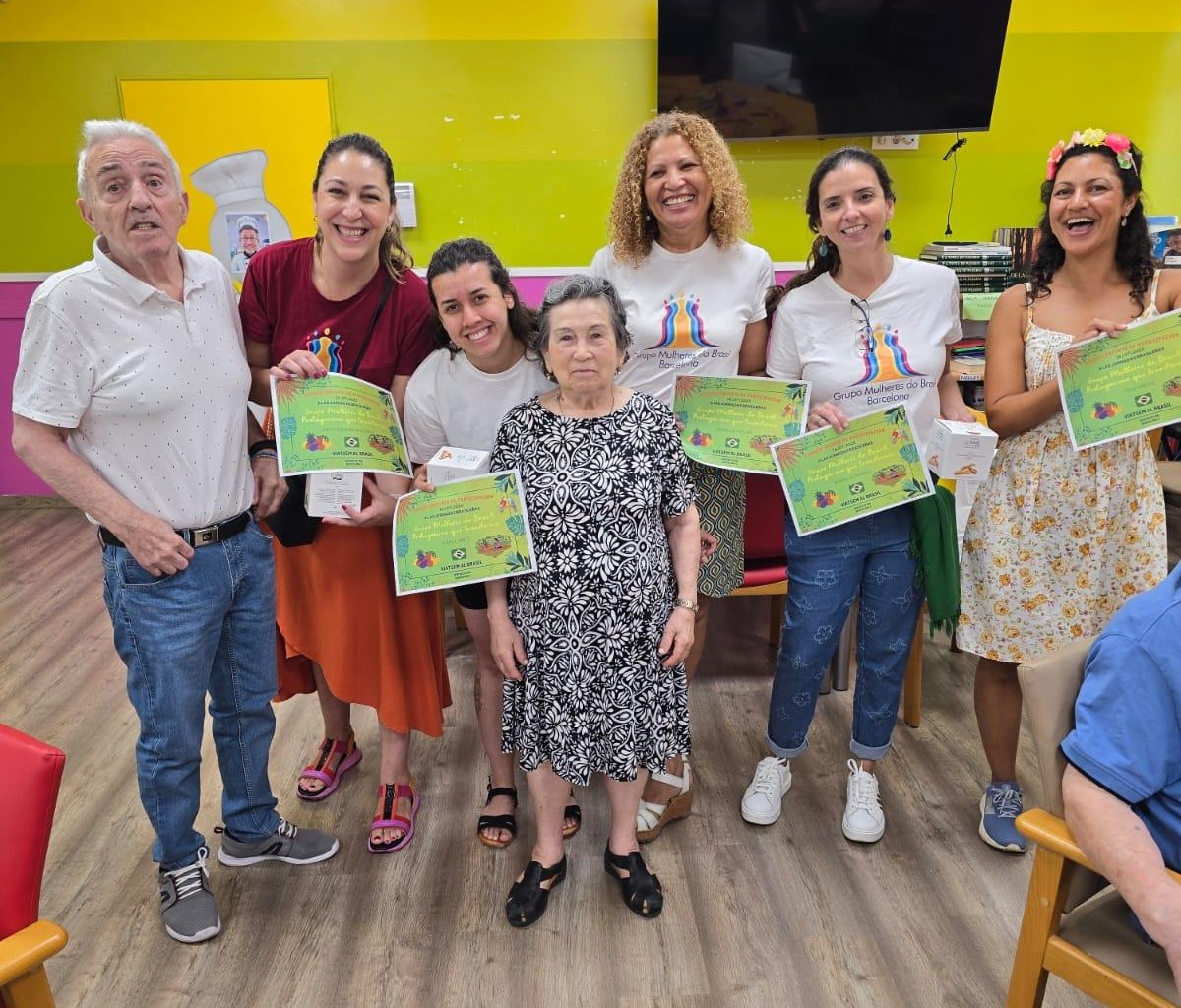 Foto de grupo em ambiente interno de um senhor e uma senhora idosos no centro, rodeados por cinco mulheres sorridentes, todas segurando certificados. O fundo é uma parede de cor verde-limão e amarela.