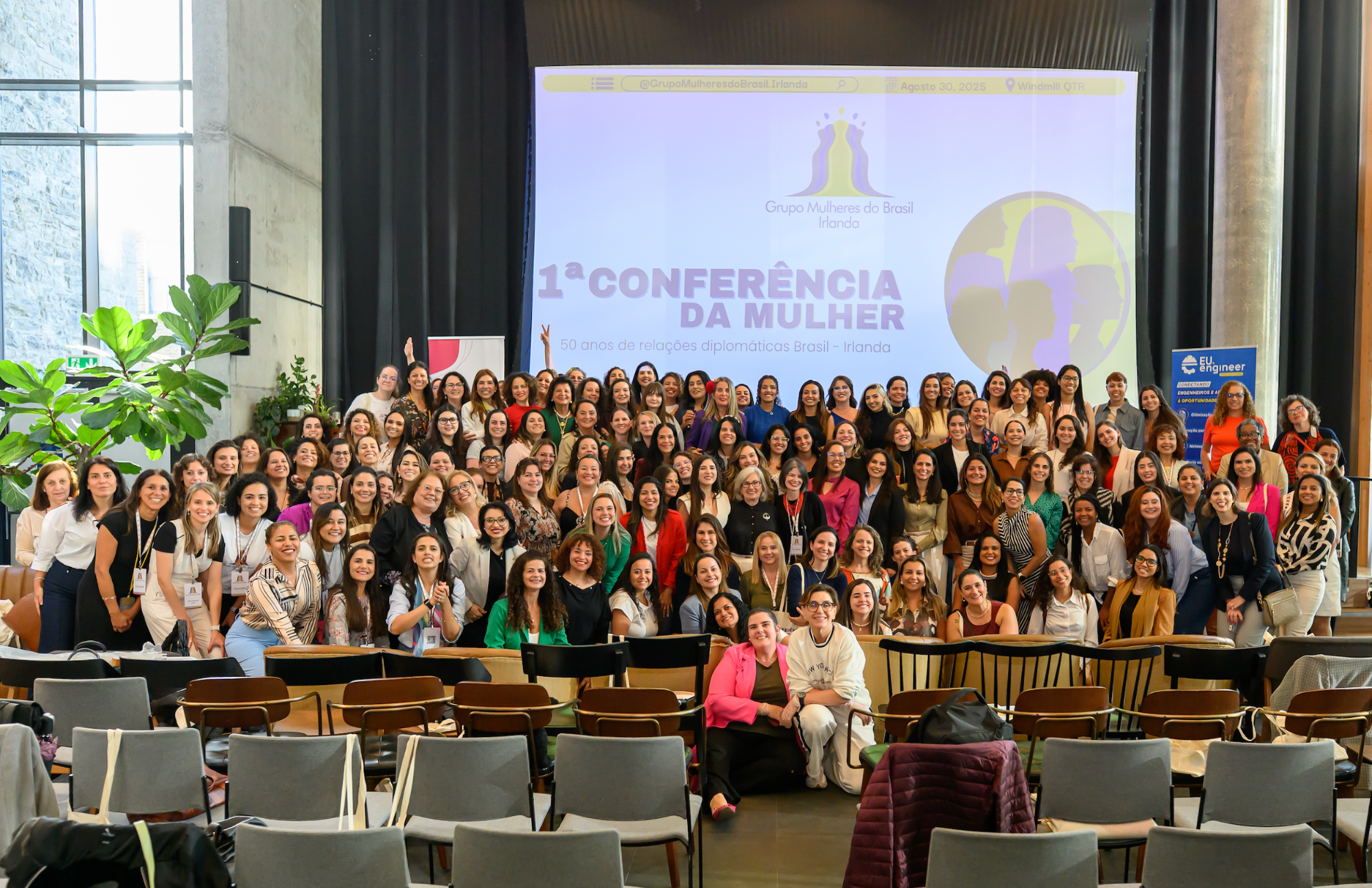 Uma grande foto de grupo com mais de cem mulheres sorrindo em um auditório. Elas estão posando em frente a uma tela grande que exibe a logomarca “Grupo Mulheres do Brasil Irlanda” e o texto “1ª CONFERÊNCIA DA MULHER”.